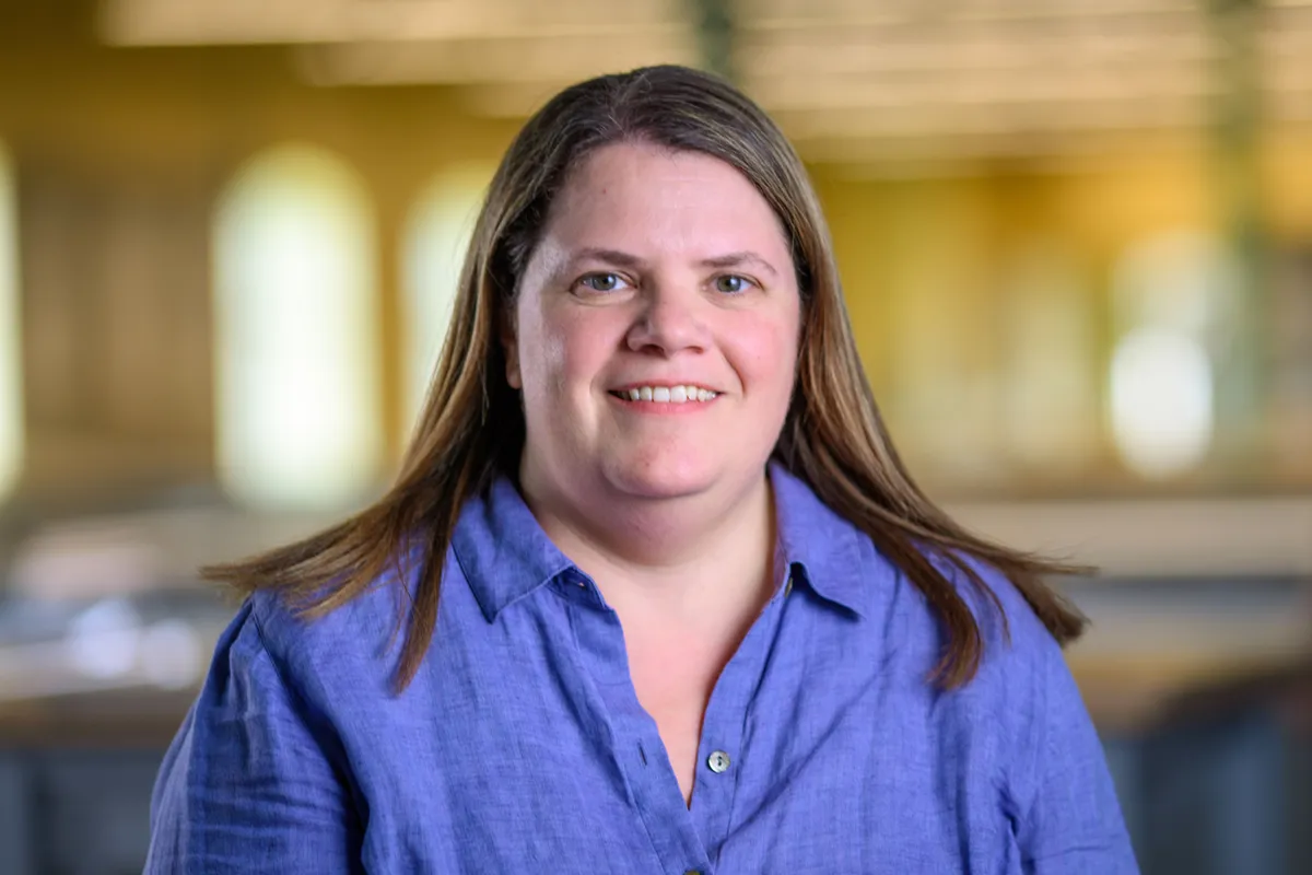 A woman with long brown hair, wearing a blue button-down shirt, smiles in a headshot with a blurred indoor background.