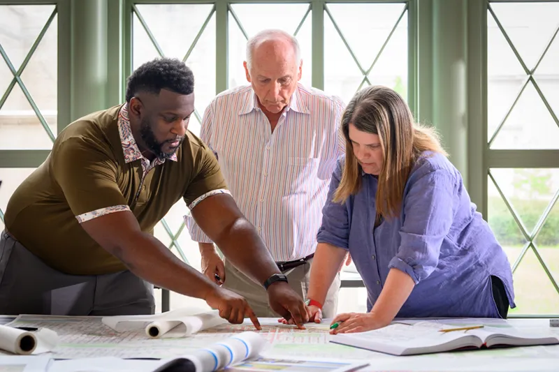 Three people, two men and a woman, lean over a table pointing at architectural plans in a well-lit room.