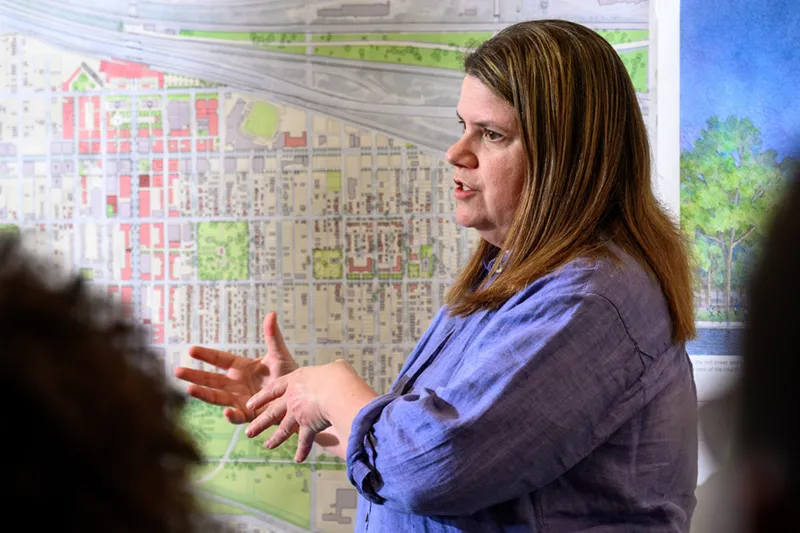 A woman in a blue shirt gestures while speaking in front of a large planning map of a city.