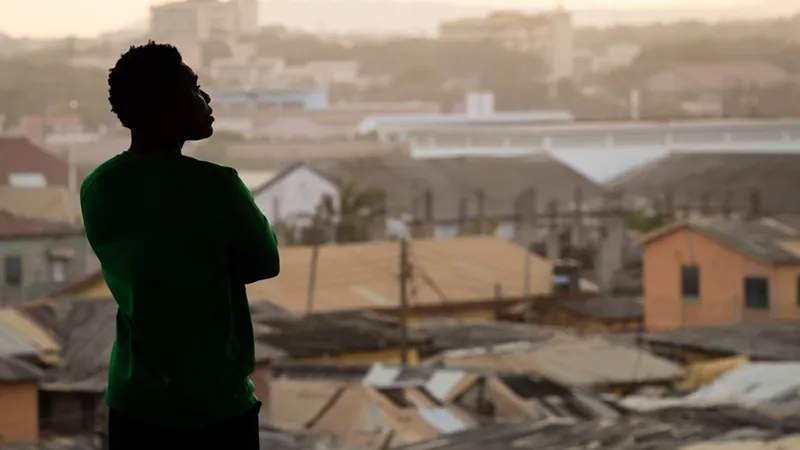 Silhouette of a person in a green shirt with arms crossed looking out over a densely built, hazy cityscape.