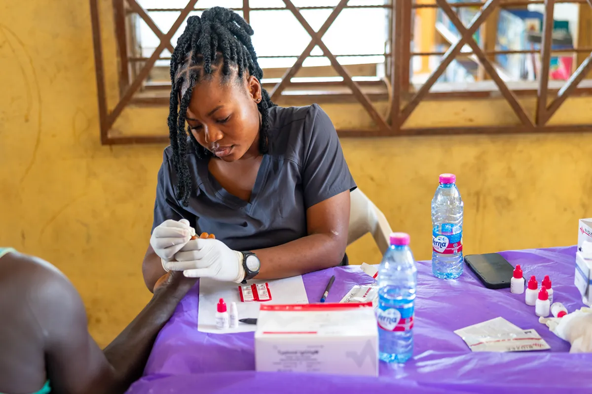 A medical worker with braided hair, wearing white gloves, performs a finger-prick blood test on a patient in a room with yellow walls and latticed windows.