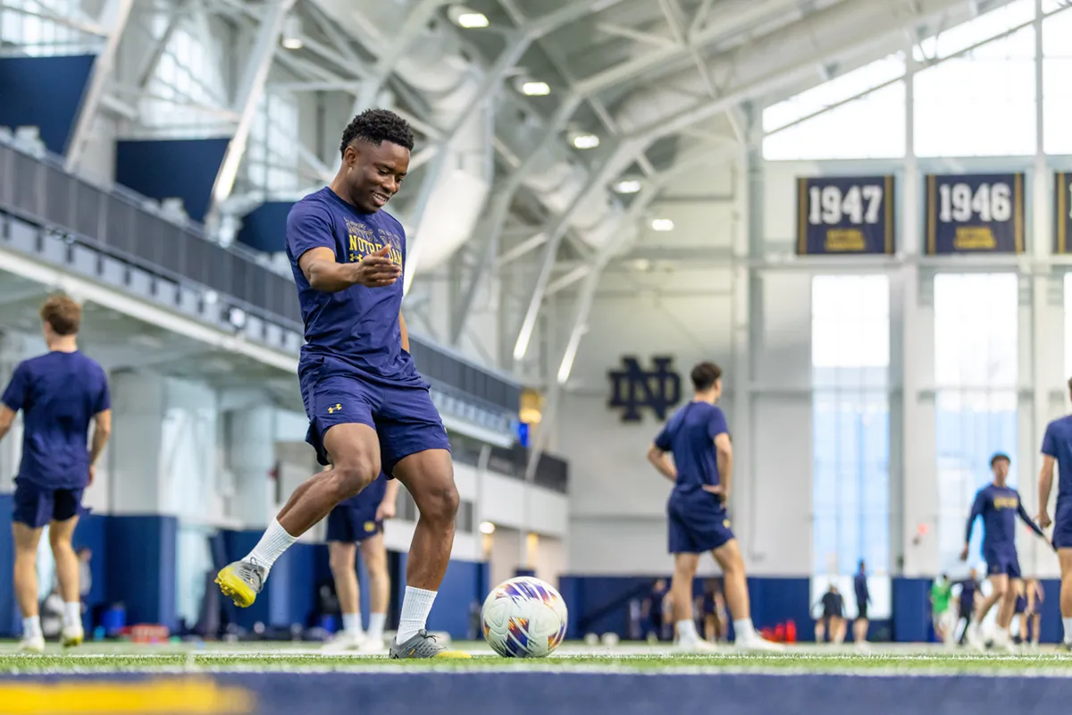 A smiling male soccer player in a navy Notre Dame uniform dribbles a ball on an indoor turf field while other players practice in the background.