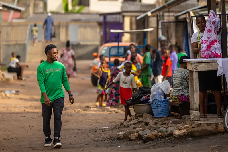 A smiling man wearing a bright green Notre Dame long-sleeve shirt walks down a dusty street filled with people and activity in a Ghanan neighborhood.