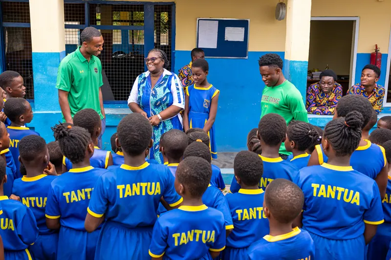 A group of children in bright blue uniforms face two smiling adults in a schoolyard setting with yellow and blue painted walls.