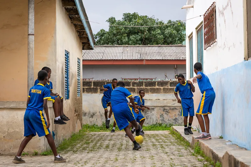 Several boys in matching blue and yellow soccer uniforms play with a yellow ball in a narrow outdoor alley between two buildings.