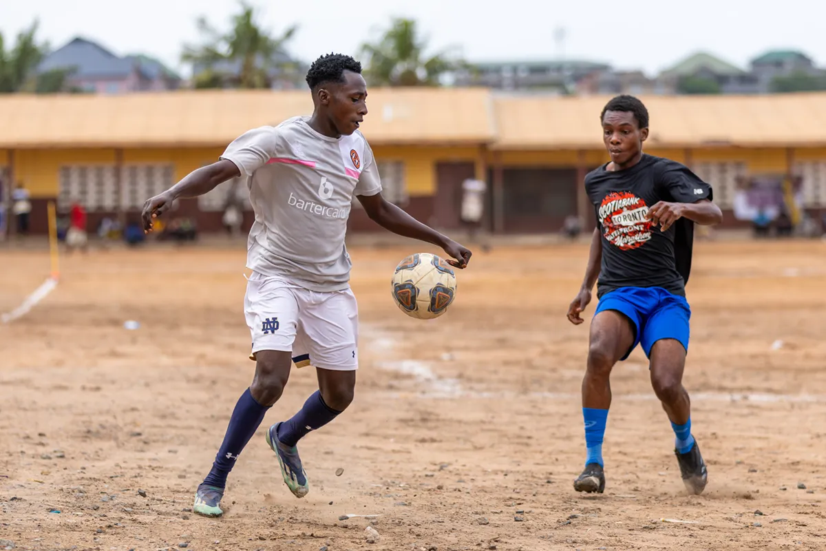 Two young men in soccer attire play on a dirt field under an overcast sky, one dribbling a ball past the other.