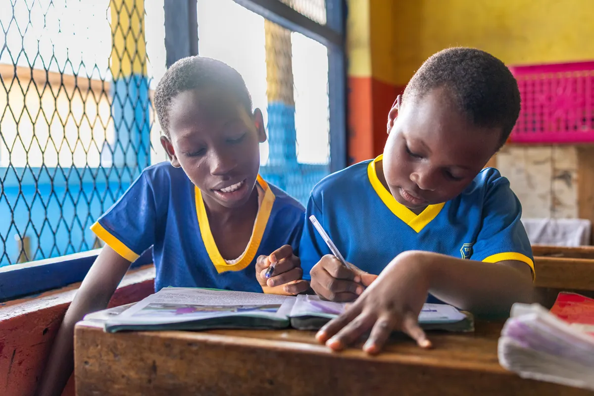 Two young boys in matching blue and yellow shirts concentrate on writing in a shared notebook at a wooden desk in a classroom.