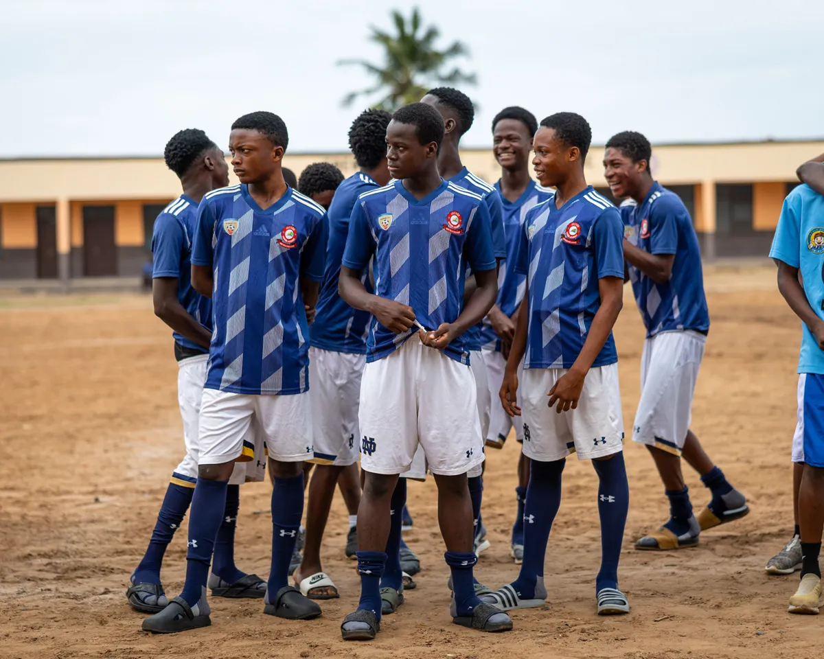 A group of young male soccer players in matching blue patterned jerseys and white shorts stand on a dirt field, some looking at the camera and others interacting.