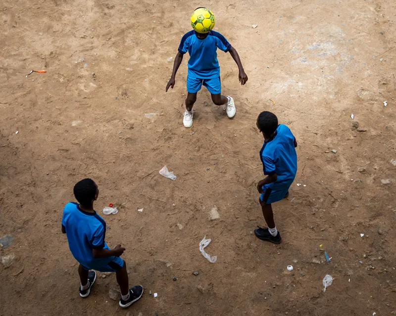 Three boys in blue soccer uniforms play with a yellow ball on a bare dirt field, viewed from a high angle.