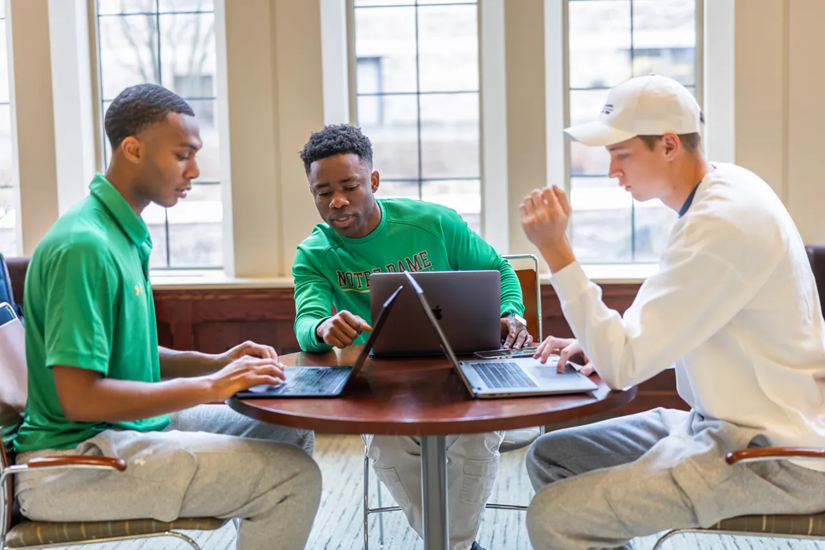 Three male students wearing Notre Dame apparel work collaboratively at a round table in a bright atrium with large windows.