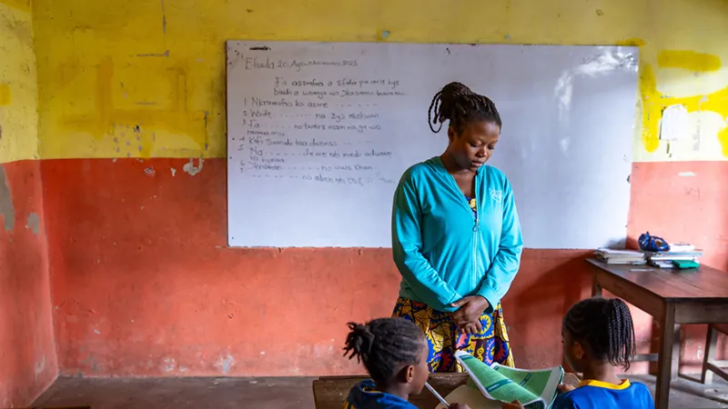 A female teacher in a bright blue jacket stands before a whiteboard covered in writing, looking down at two young students in a classroom with yellow and red walls.