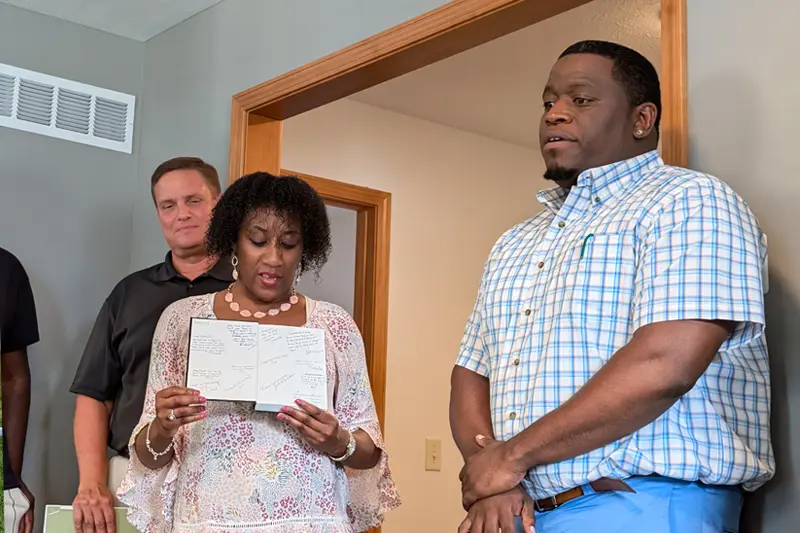 A woman reads aloud from a notebook filled with handwritten messages.	A man stands listening behind her to her left, and another man stands to her right with his hands clasped. They appear to be inside a house or office.