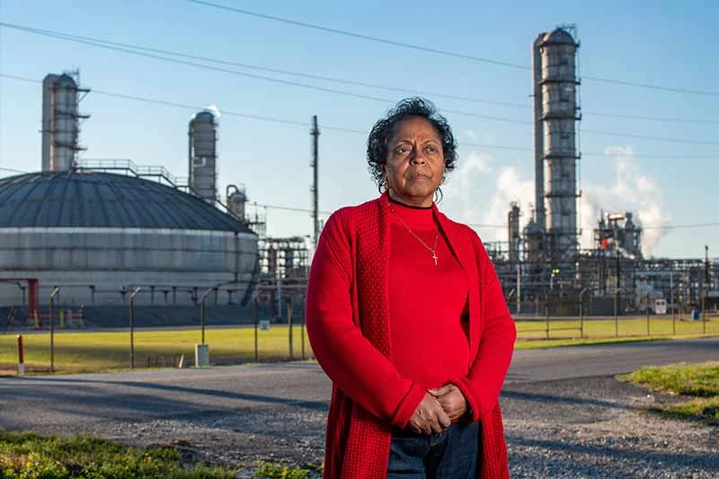 A woman in a red sweater stands with her hands clasped in front of an industrial plant.