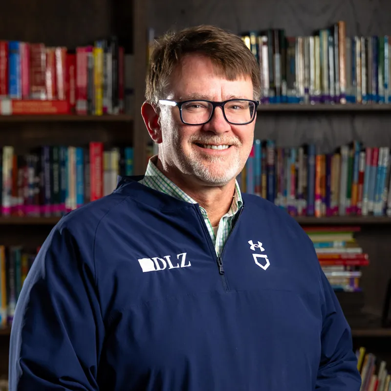 Smiling middle-aged man with short brown hair, a beard, and rectangular glasses wearing a navy DLZ quarter-zip pullover over a plaid shirt, with bookshelves visible in the background.