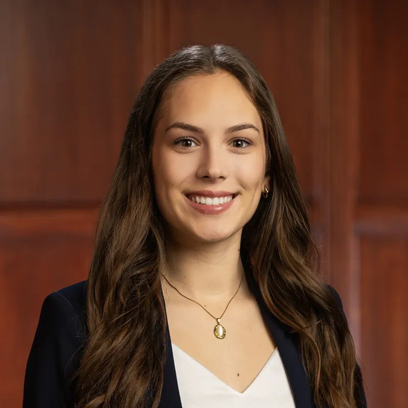 Smiling young woman with long brown hair wearing a navy blazer, white top, and a small gold oval pendant necklace, photographed against a warm wood-paneled background.