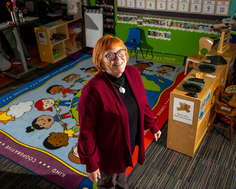 Smiling woman with short red hair and dark-framed glasses wearing a burgundy cardigan and black turtleneck, standing in a colorful early childhood classroom with a children's world map rug and learning materials behind her.