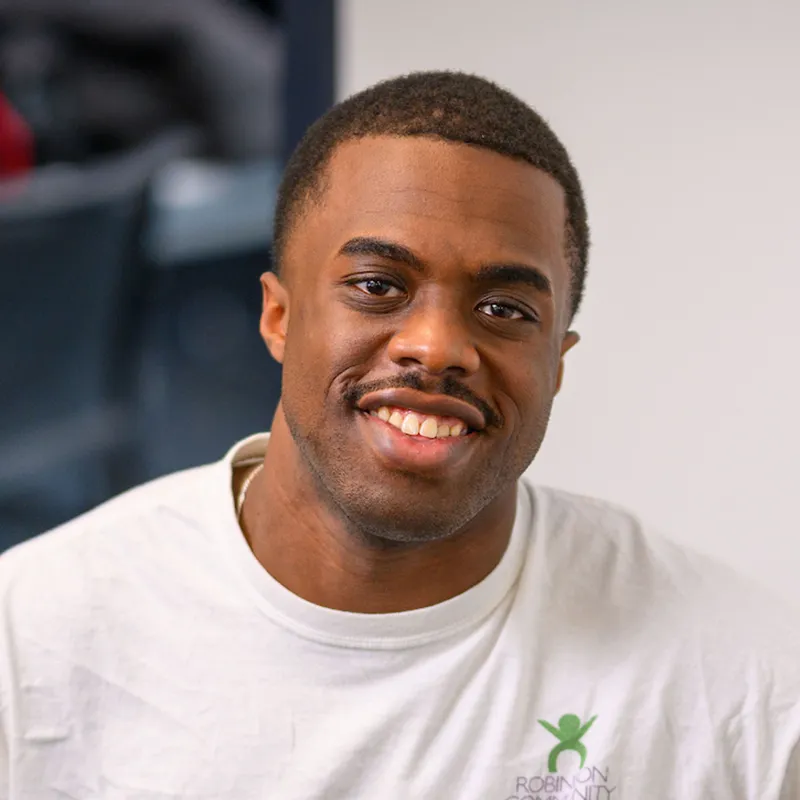 Smiling young man with short hair wearing a white t-shirt with a green Robinson Community Center logo, photographed against a neutral blurred background.