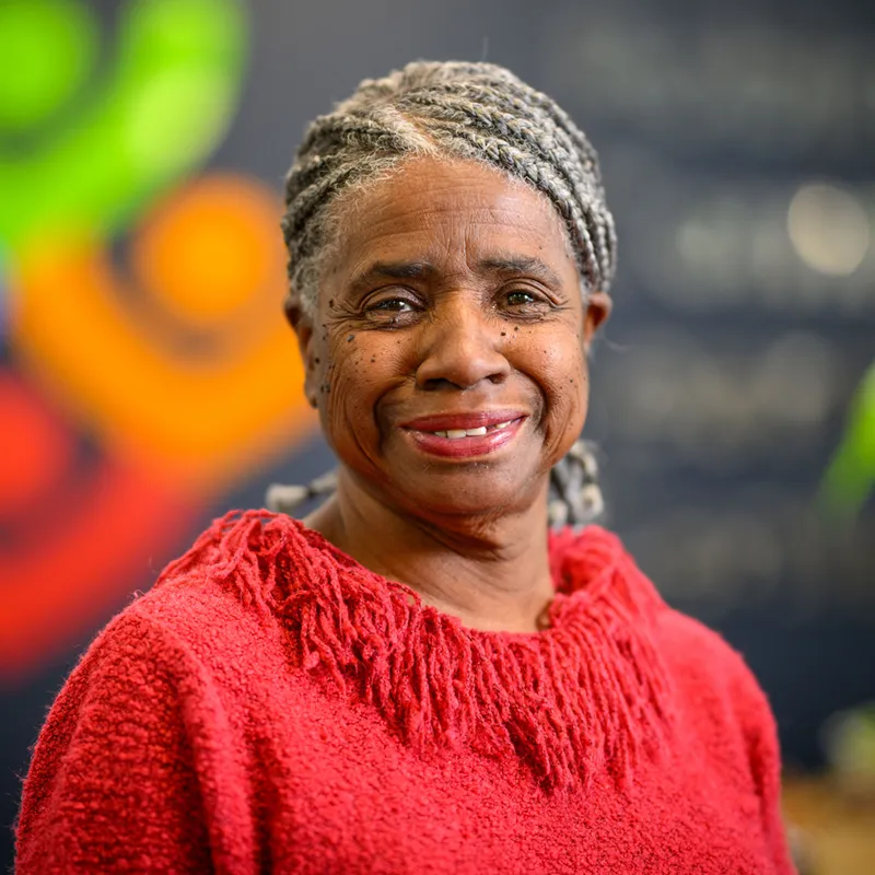 Smiling older woman with gray braided hair wearing a red fringed sweater, photographed against a blurred colorful background with green and orange shapes.
