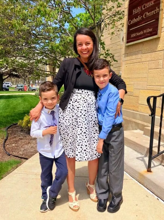 A smiling woman in a black blazer and patterned skirt poses with her arms around two dressed-up young boys outside Holy Cross Catholic Church in South Bend, Indiana.