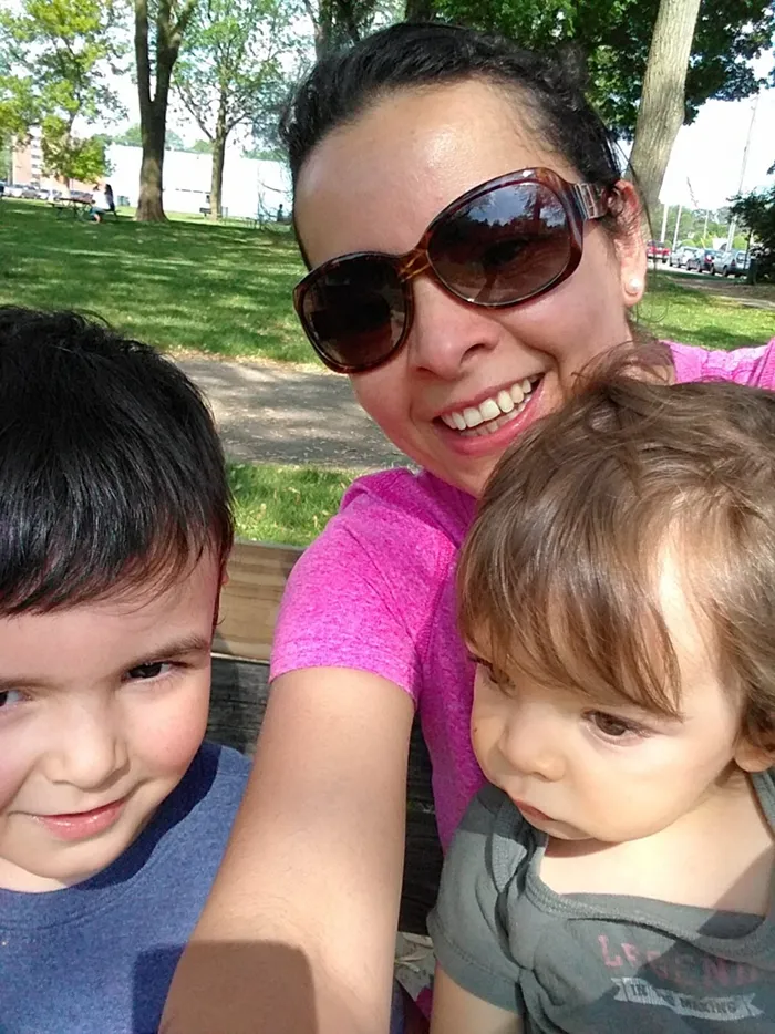A smiling woman wearing large sunglasses and a pink shirt takes an outdoor selfie with two young children at a park picnic table on a sunny day.