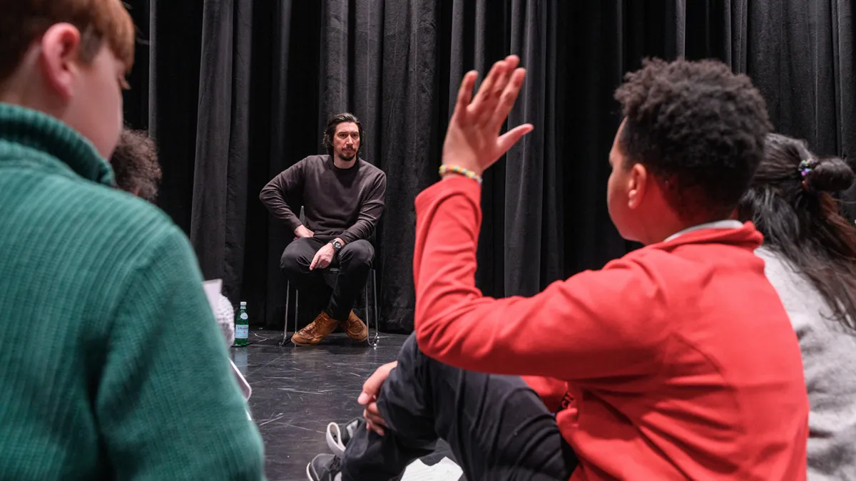 Adam Driver in a dark sweater sits on a chair on a black box theater stage, listening attentively as a child in a red jacket raises their hand to ask a question during a Q&A session.