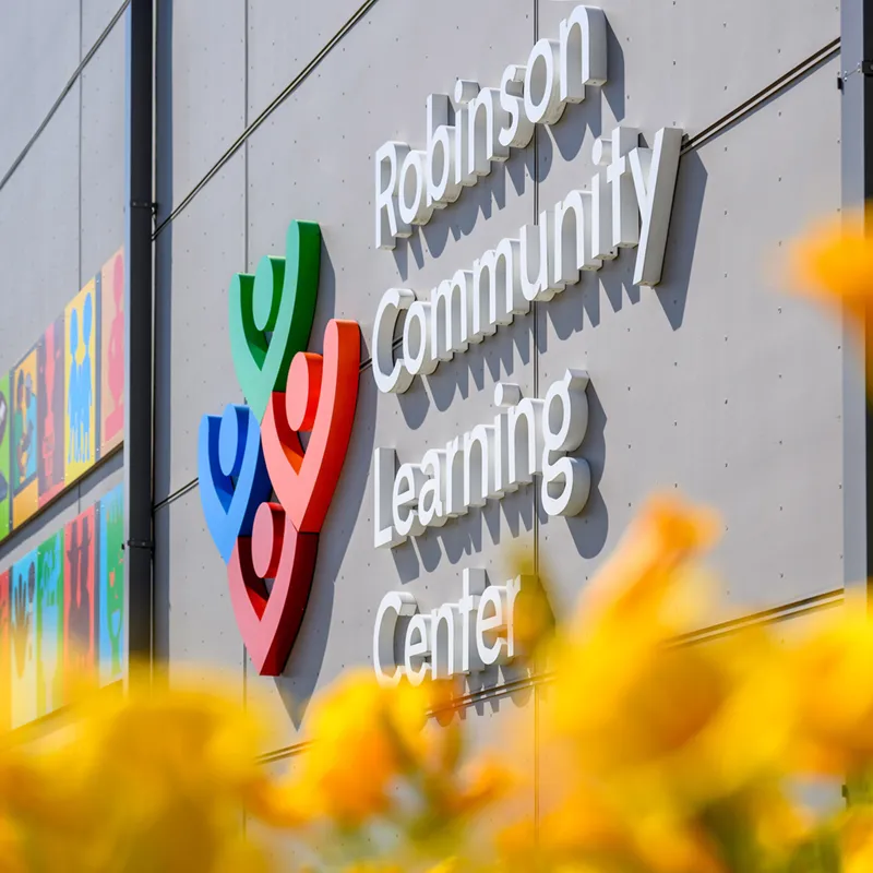 Exterior façade of the Robinson Community Learning Center featuring the organization's colorful logo and white dimensional lettering, with yellow flowers blurred in the foreground and a vibrant mural visible along the left edge.