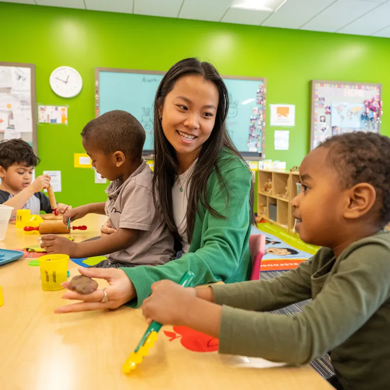 A smiling young woman in a green cardigan engages with preschool children during a Play-Doh activity at a classroom table, with a bright green wall and learning materials visible behind them.