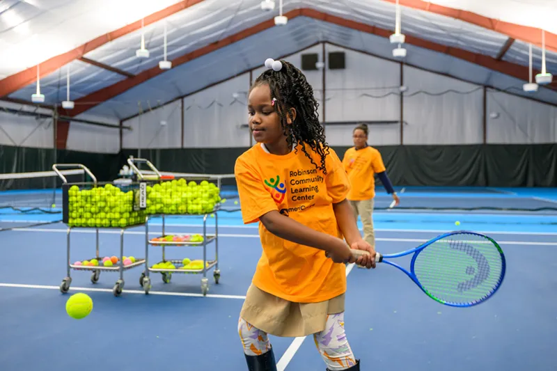 A young girl with braided hair holds a tennis racket at the net on an indoor court, wearing a yellow Robinson Community Learning Center AmeriCorps t-shirt, with other players visible in the background.