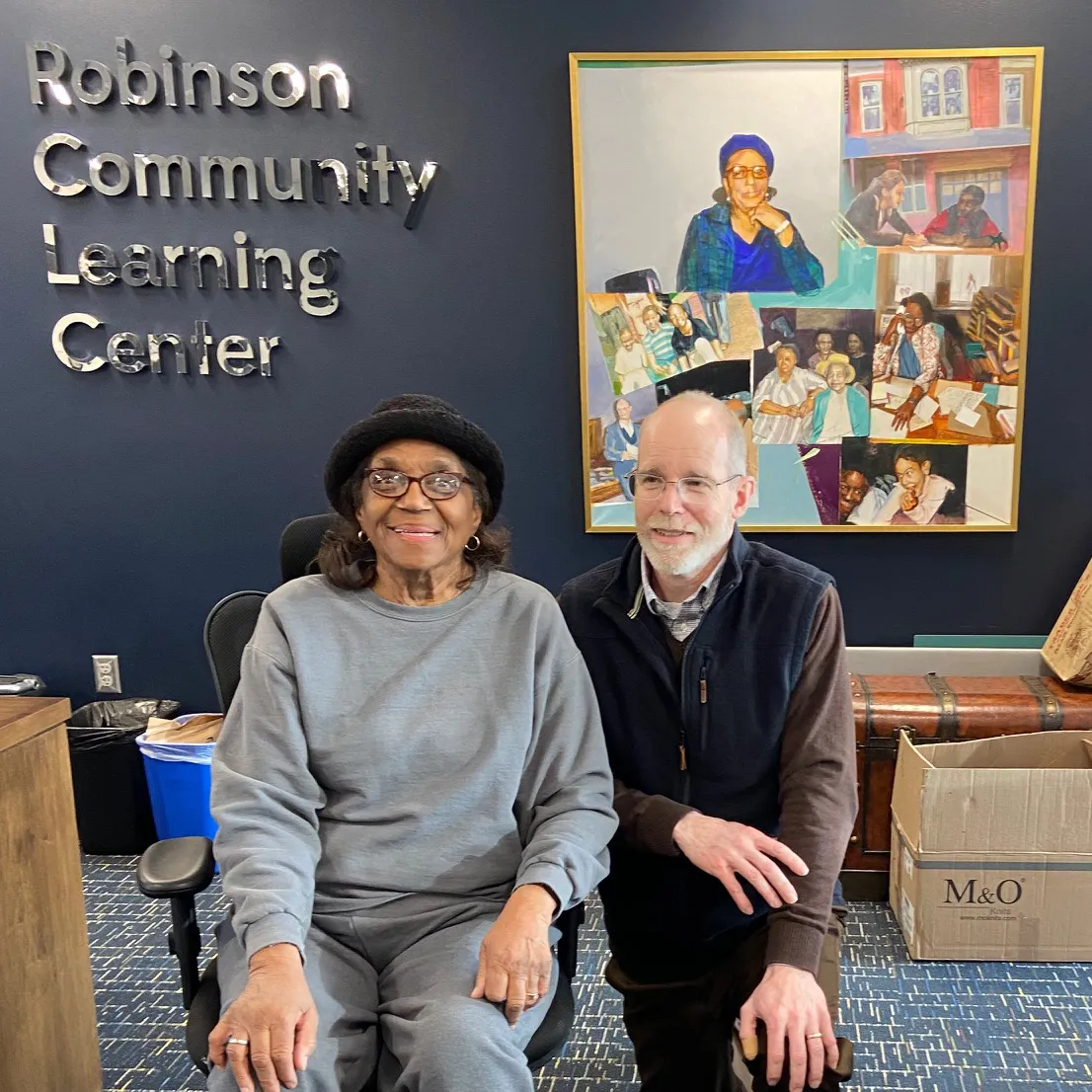 Two smiling people seated together inside the Robinson Community Learning Center, with the organization's name on the dark blue wall behind them and a large Renelda Robinson portrait collage displayed above.
