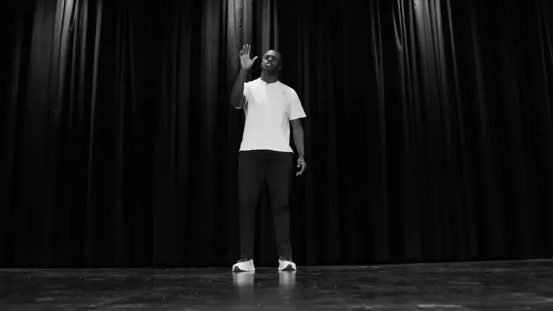 Black-and-white image of a young man in a white t-shirt, dark pants, and white sneakers standing alone on a stage, one hand raised, against a backdrop of dark curtains.
