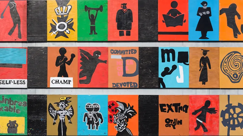 A colorful community mural made of individual painted panels featuring bold black silhouette figures and words including 'Self-Less,' 'Champ,' 'Committed,' 'Devoted,' 'Unbreakable,' and 'Extraordinary' on vibrant red, orange, blue, and green backgrounds.