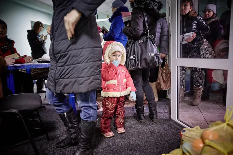 A small Ukrainian child in a red winter coat with cream fur, red pants with gold butterflies, and light blue gloves stands looking forward, sucking a gloved thumb. Adults in winter clothes fill a crowded indoor waiting area, with others reflected in a glass door. Bags are on the floor.