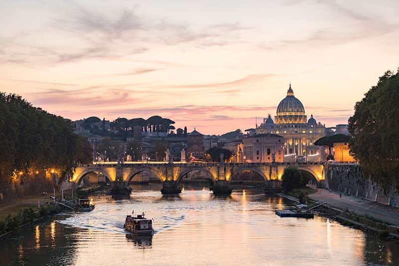 Sun sets over the city of Rome. In the foreground is the Sant'Angelo bridge which spans the Tiber River. In the background to the right is the dome of	Saint Peter's Basilica.