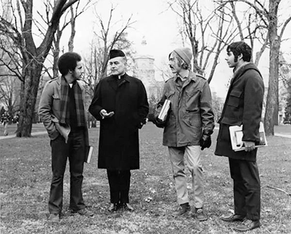 Father Theodore Hesburgh stands on a grassy campus lawn talking with three male students. He is wearing a dark overcoat and clerical collar. The students hold books, and the Main Building is visible in the distance.