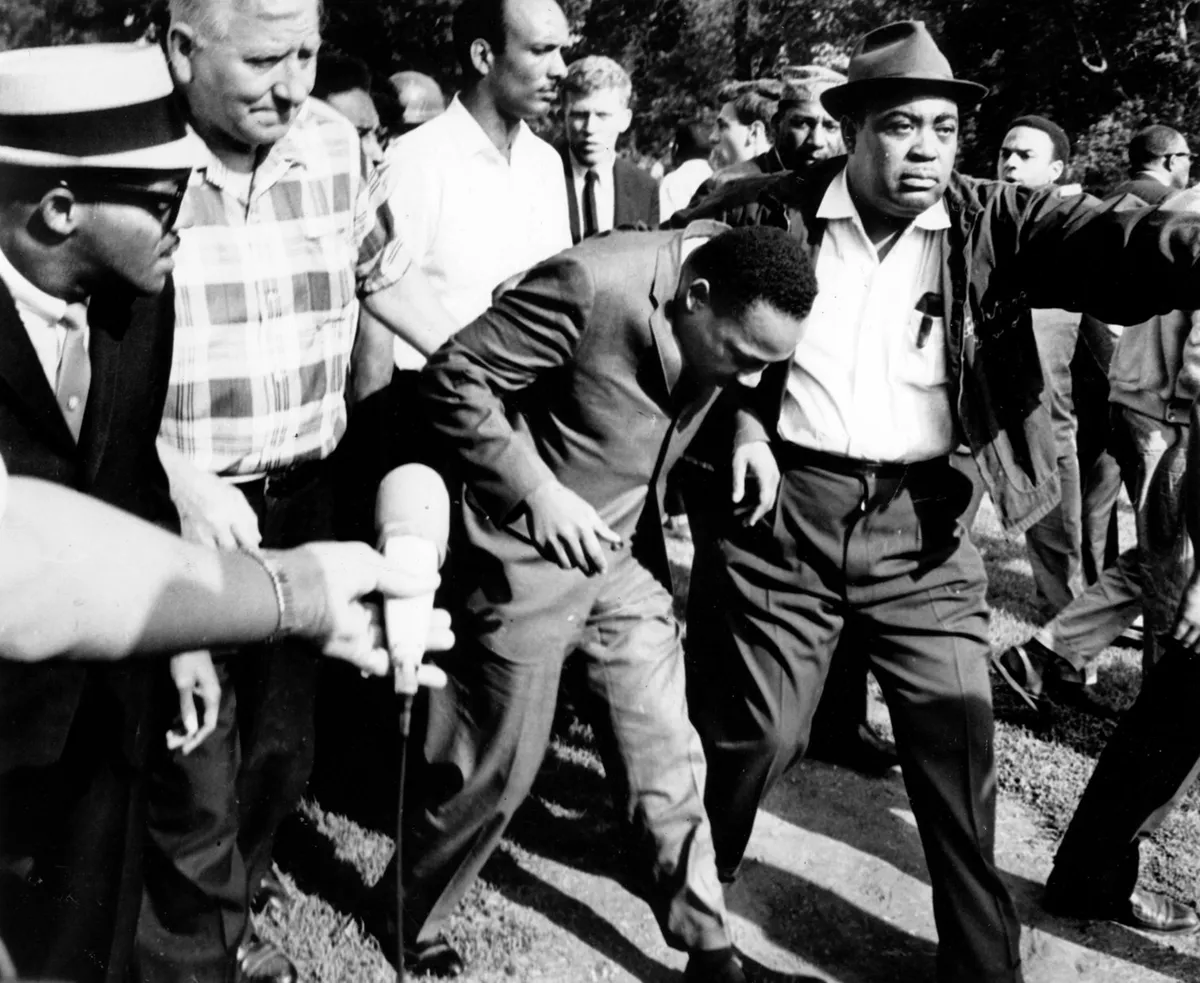 Dr. Martin Luther King Jr. ducks in a suit as a man in a fedora reaches out to shield him during a chaotic outdoor moment. Anson can be seen in the background facing the camera.