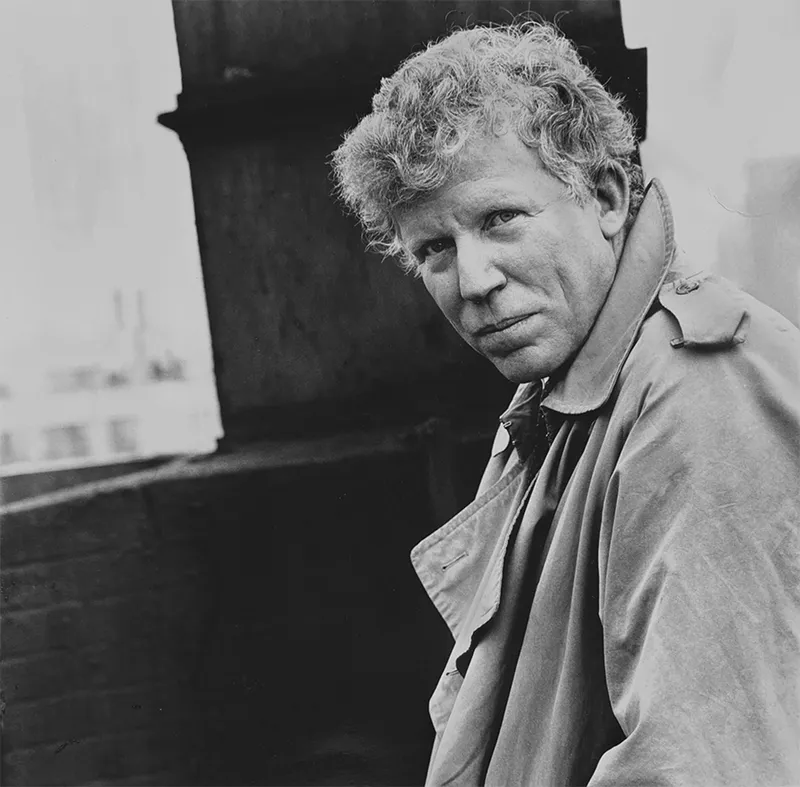Robert Sam Anson looks over his shoulder with a neutral expression. He has light, curly hair and is wearing a tan trench coat with the collar turned up. The black and white photo has a shallow depth of field with a dark, blurred background.