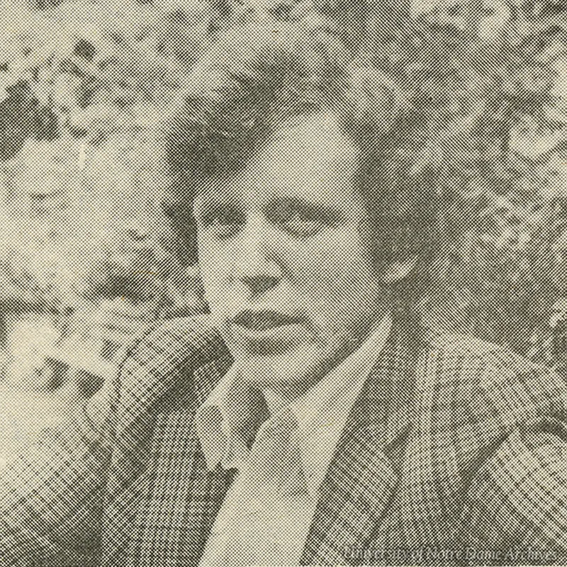 A black and white grainy photo shows a man with wavy hair wearing a patterned blazer over a collared shirt. He is looking toward the camera with a neutral expression against a blurred outdoor background.