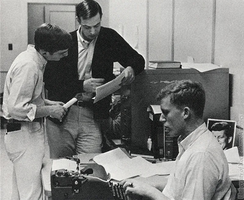 Black and white image of three men in an office. One man in the foreground types on a typewriter, while two men in the background stand and review a document together. A small framed portrait of John F. Kennedy sits on the desk.