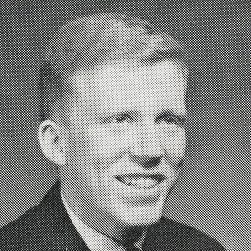 A black and white yearbook portrait of a smiling young man with short, light-colored hair. He is wearing a dark suit jacket and a white collared shirt. The image has a visible halftone dot pattern.