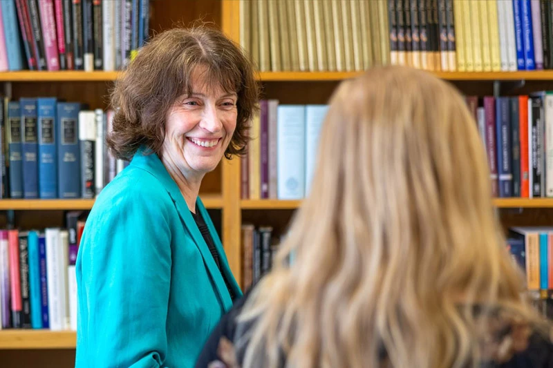 A smiling woman with wavy brown hair in a teal blazer looks towards a blonde-haired person, with bookshelves behind her.