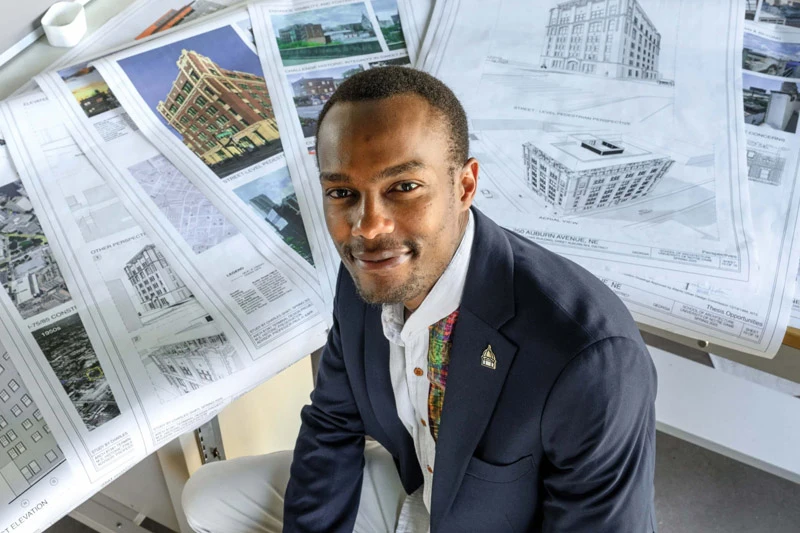 A smiling man in a dark suit, colorful patterned shirt, and a lapel pin looks up from a table covered in architectural drawings. The drawings feature detailed building designs, including one prominent red brick building.