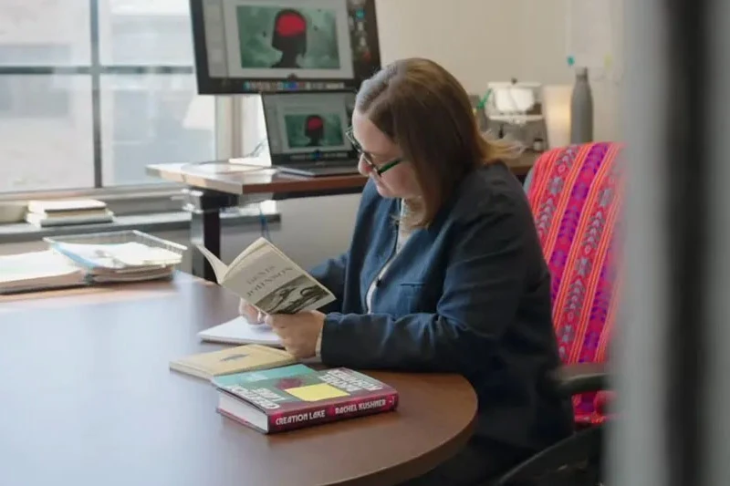 A woman in green glasses and a dark blazer reads an open book at a brown desk. Several books are on the desk. Behind her, two computer monitors display a dark head silhouette with a red brain section.