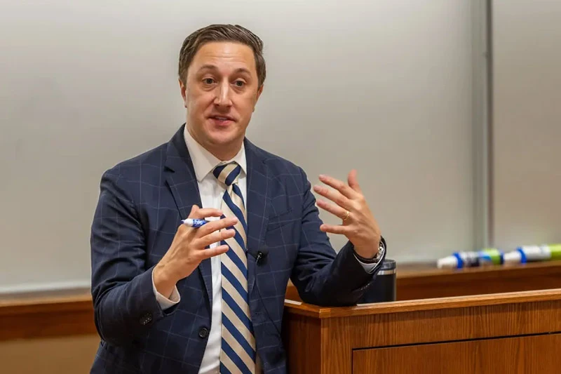 A man in a blue windowpane suit and striped tie speaks expressively while leaning on a wooden lectern in a classroom.