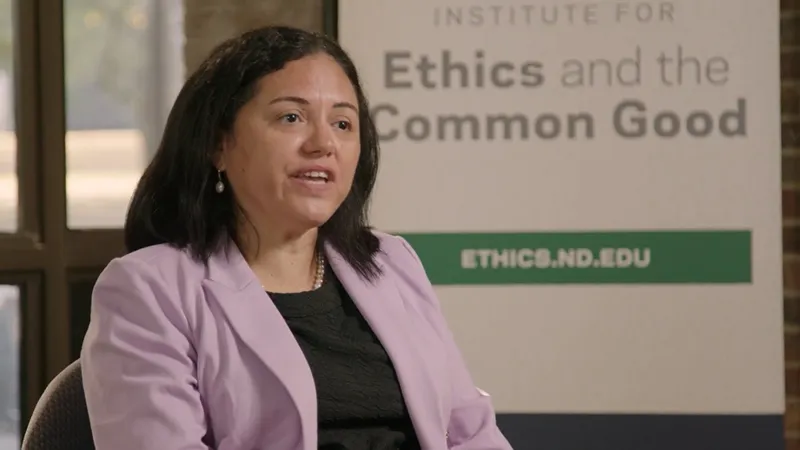 A woman with dark wavy hair speaks while wearing a light purple blazer and a pearl necklace. She sits in front of a white sign for the Institute for Ethics and the Common Good with the website ETHICS.ND.EDU visible on a green banner.
