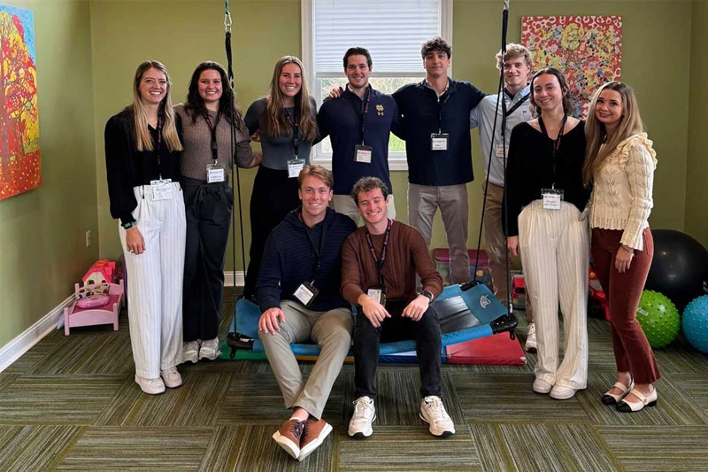 Ten smiling Notre Dame students, six women and four men, pose in a therapy room. Two men sit on a colorful swing, while eight students stand behind them. One student wears a navy polo with a gold Notre Dame logo.