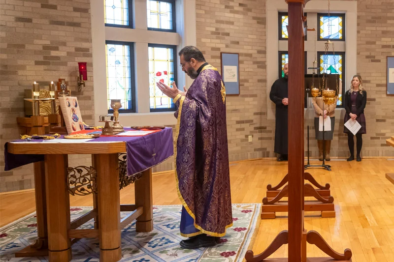 A priest in a rich purple vestment stands before an altar with lit candles, reciting prayers with his hands raised near stained-glass windows.