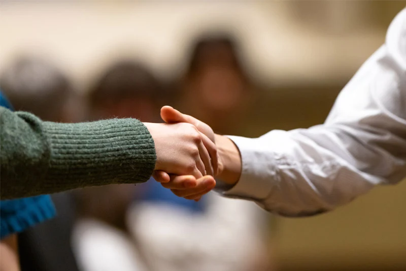 Two people shake hands. One wears a dark green sweater; the other, a white striped shirt.