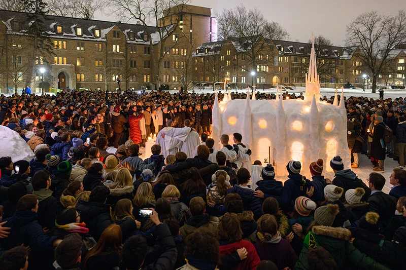 Hundreds of people, many holding candles, gather on a snowy winter night around a brightly illuminated white snow chapel with spires. Notre Dame dormitories glow in the background.