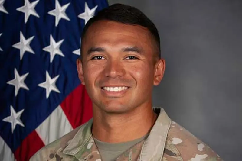 A smiling man in a U.S. Army OCP uniform stands before an American flag. 