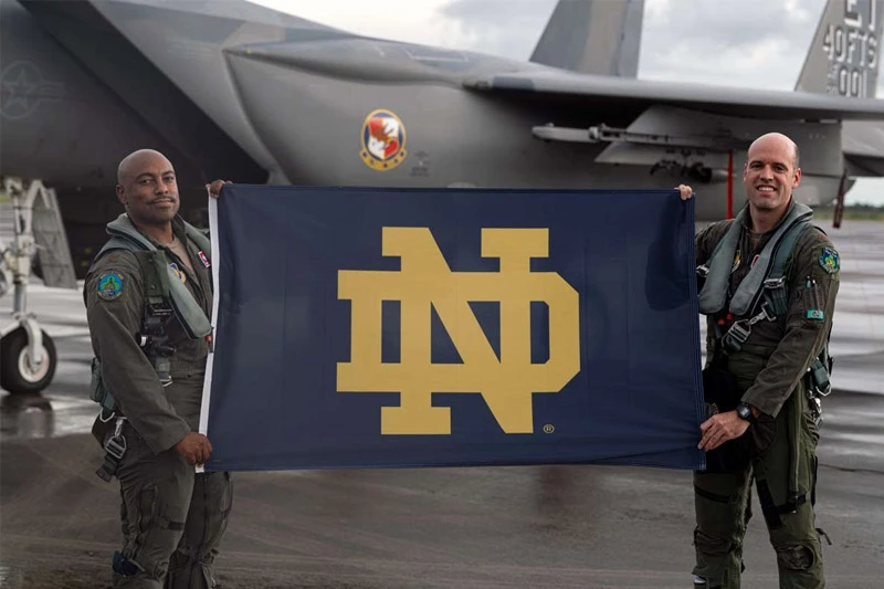 Two smiling pilots in olive flight suits hold a large navy blue Notre Dame flag with a gold ND logo, standing on a tarmac with a gray military jet in the background.